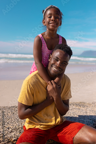Portrait of smiling african american girl embracing young father sitting at beach against blue sky
