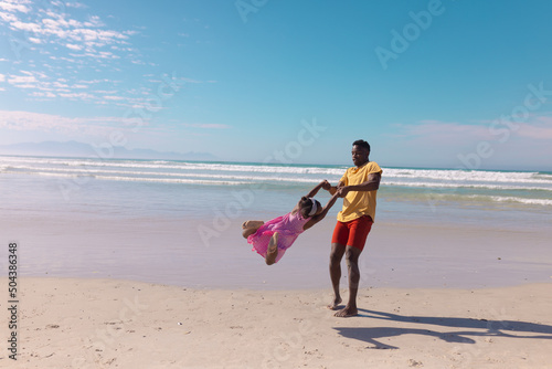 African american young man holding playful daughter's hands and spinning her at beach against sky