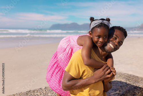 African american daughter embracing cheerful young father sitting at beach against sky on sunny day