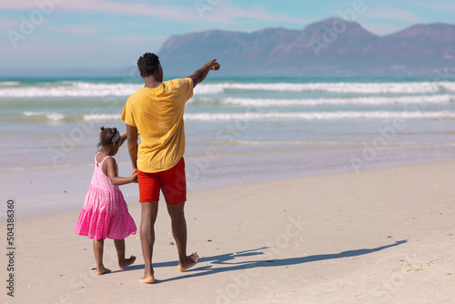 Rear view of african american young man pointing while walking with daughter at beach on sunny day