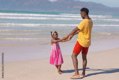 African american girl holding young father's hands while enjoying at beach against sea on sunny day