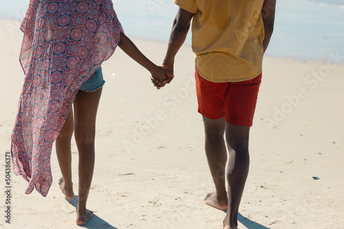 Rear view low section of african american young couple holding hands while walking on sand at beach