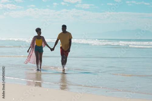 Happy african american young couple holding hands and walking in sea against sky during sunny day
