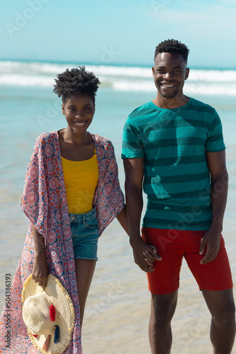 Portrait of smiling african american young couple holding hands and standing against sea and sky