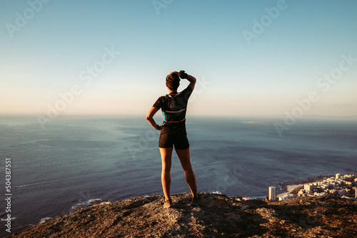 Back view of fit woman standing on cliff. Young trail runner taking a break enjoying the view at sunset.