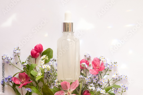 Cosmetic glass transparent bottle with dropper for hyaluronic acid among different wildflowers on a white background. Beautiful light reflections. Mockup bottle