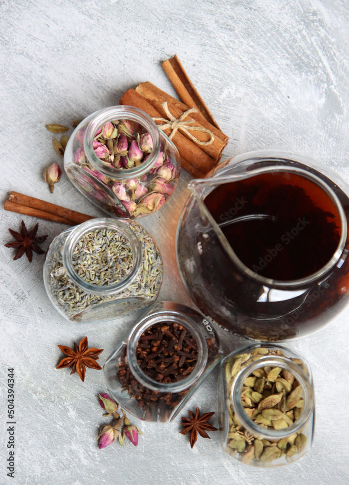 Fototapeta premium Teapot and spices jars on light grey background with copy space. Different tea flavors on a table. Lavender, cardamom, cinnamon, rose, anise stars and clove top view photo. 
