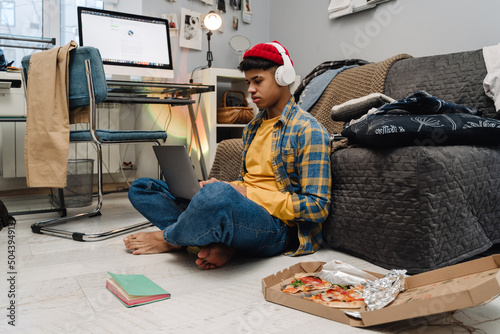 Teenage boy using laptop and eating pizza while studying at home