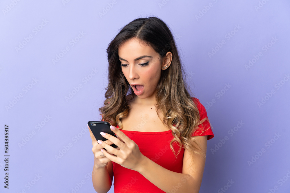 Young caucasian woman isolated on purple background looking at the camera while using the mobile with surprised expression Young caucasian woman isolated on purple background looking at the camera while using the mobile with surprised expression