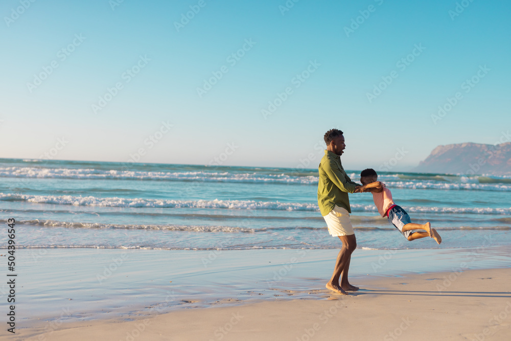 Playful african american young father spinning son at beach against clear sky during sunset