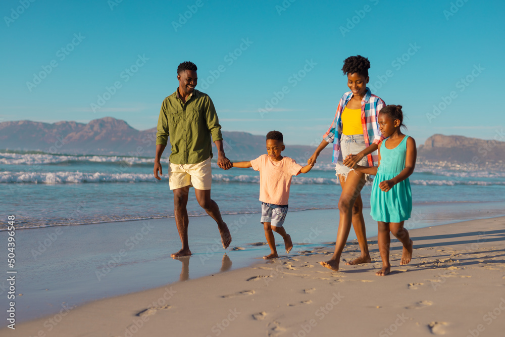 Happy african american young parents holding children's hands and walking at beach against clear sky