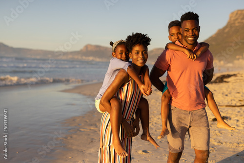Portrait of happy african american young parents piggybacking son and daughter at beach against sky
