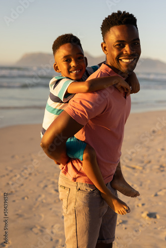Portrait of smiling african american young man piggybacking son at beach against clear sky at sunset