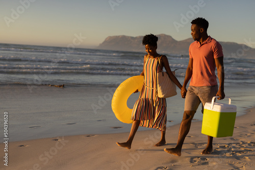 African american young couple with cooler and inflatable ring holding hands while walking on beach