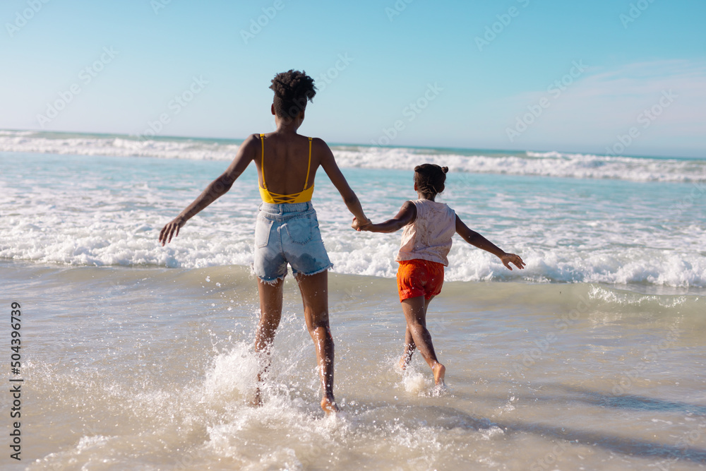Rear view of african american young mother holding girl's hands and enjoying in sea against sky