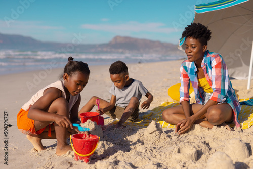 African american young mother sitting with son and daughter playing with sand at beach against sky