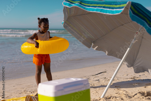 African american girl wearing swim ring while standing by umbrella at beach against sky on sunny day