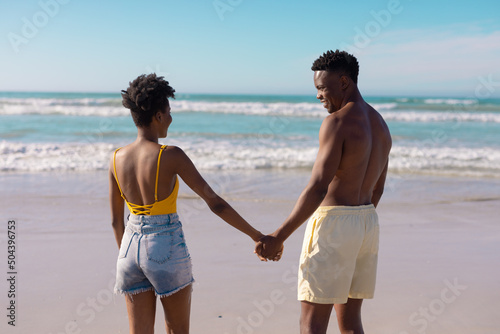 African american shirtless young man holding woman's hands while standing at beach against sky