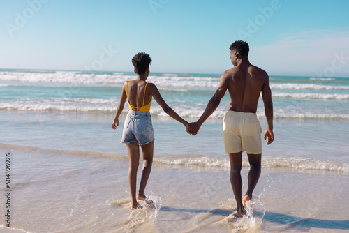 Rear view of african american young couple holding hands and walking towards sea against sky