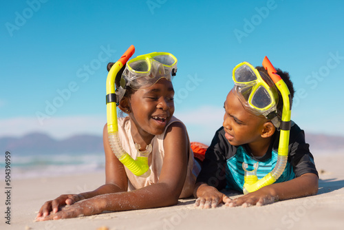 African american boy and girl wearing snorkels and masks talking while lying on sand against sky