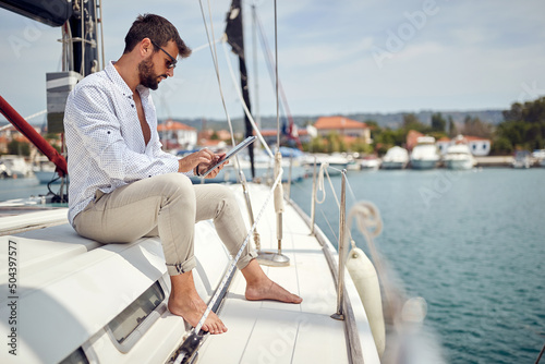 A young man is sitting on a yacht and using a tablet while riding through the dock on the seaside. Summer, sea, vacation