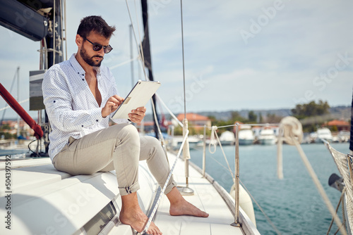 A young businessman is sitting on his yacht and doing some job on his tablet while riding in the dock on the seaside. Summer, sea, vacation © luckybusiness