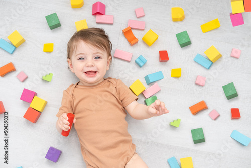 cute smiling baby girl in brown bodysuit laying on floor around colorful wooden blocks. top view photo