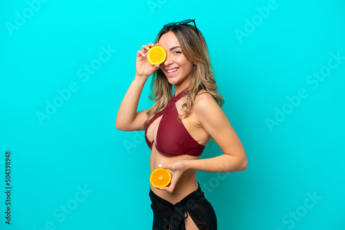 Young caucasian woman isolated on blue background in swimsuit and holding an orange