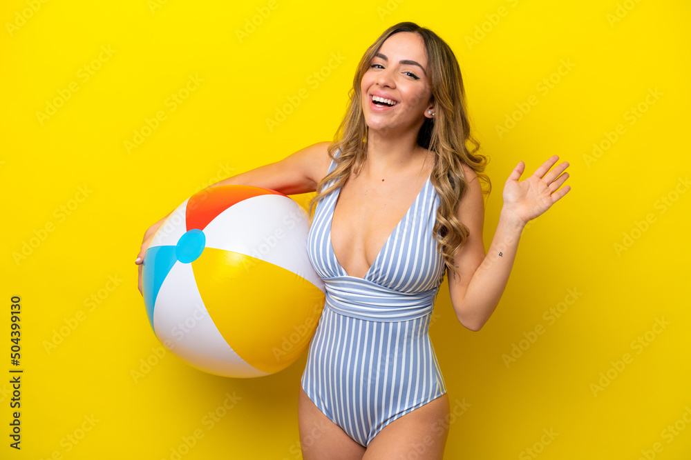 Young woman holding beach ball isolated on yellow background saluting with hand with happy expression Young woman holding beach ball isolated on yellow background saluting with hand with happy expression