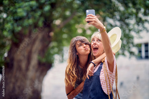 Young women making funny and goofy face expressions and taking selfie, on street. Young women walking on street of European city. Travel, fun, togetherness concept. photo