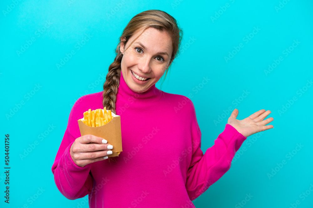 Young caucasian woman holding fried chips isolated on blue background extending hands to the side for inviting to come Young caucasian woman holding fried chips isolated on blue background extending hands to the side for inviting to come