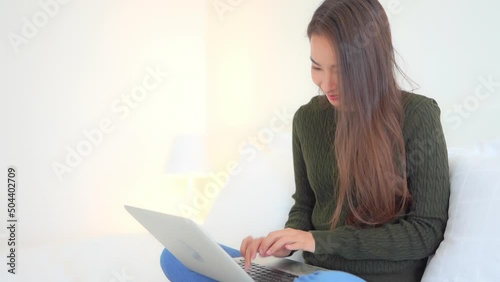 Close-up of a casually dressed young woman sitting on a bed as she works on her laptop. Title space photo