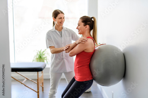 Physiotherapist helping woman do wall squats with fit ball. Young female patient doing back exercise using fitball in physio room of modern clinic