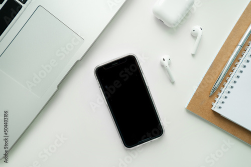 Mobile phone with blank black screen on desktop. Business concept template: laptop, mobile phone, wireless headphones, notepad and pen.