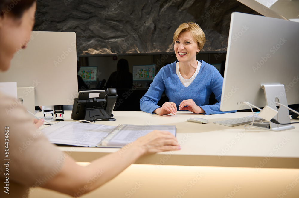 Beautiful pleasant Caucasian female travel agent talking to her client, showing catalog with tours or consumer services while sitting in modern office
