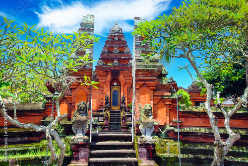 Beautiful red hindu temple gate, green trees, blue sky, mythological figures - Pura Batuan, Ubud, Bali photo