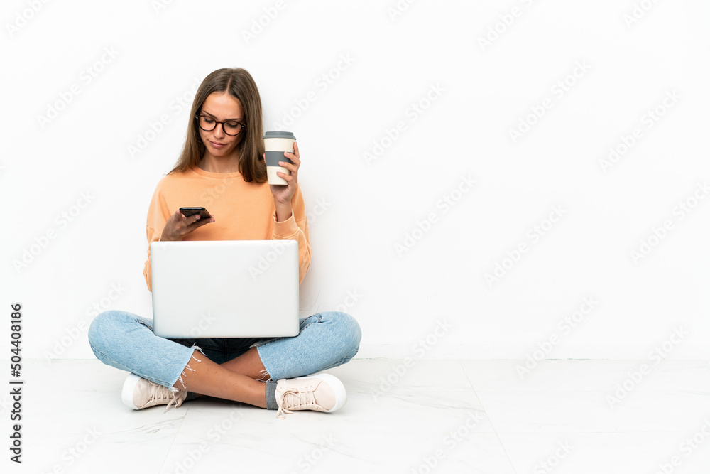 Young woman with a laptop sitting on the floor holding coffee to take away and a mobile Young woman with a laptop sitting on the floor holding coffee to take away and a mobile