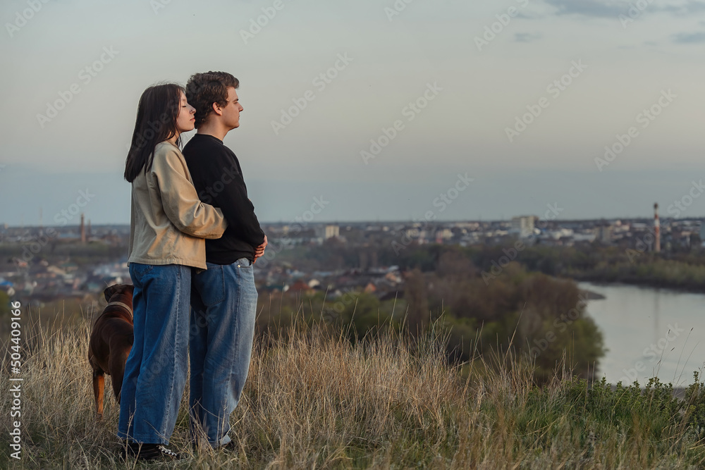 a guy and a girl in love, embracing, look at a beautiful view of nature in front of them, their big dog is with them in the warm season