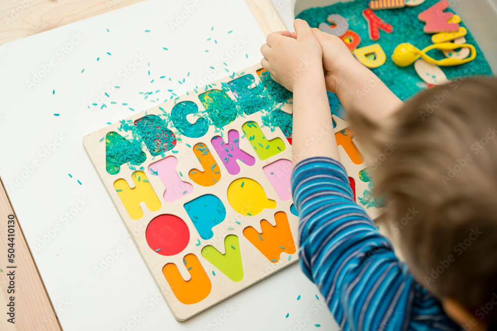 kid learning to read alphabet through game. Sensory activity with colored rice and wooden letters. Educations at home, pre-school education, Montessori methodology kid learning to read alphabet through game. Sensory activity with colored rice and wooden letters. Educations at home, pre-school education, Montessori methodology