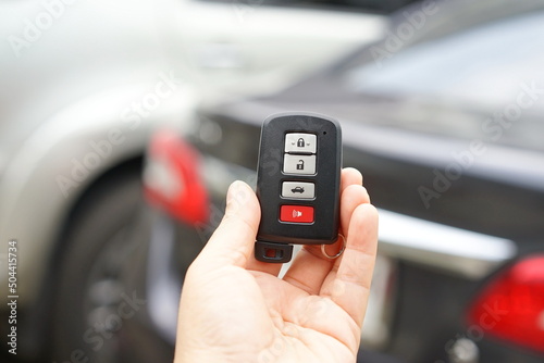 Left hand holding a black remote car key. There is a small ring hanging on it and three gray buttons for locking and unlocking the car. red button for emergency Smart key systems for modern cars 