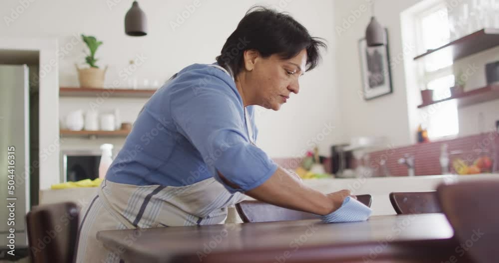 Senior biracial woman wearing apron and cleaning table in kitchen alone Senior biracial woman wearing apron and cleaning table in kitchen alone