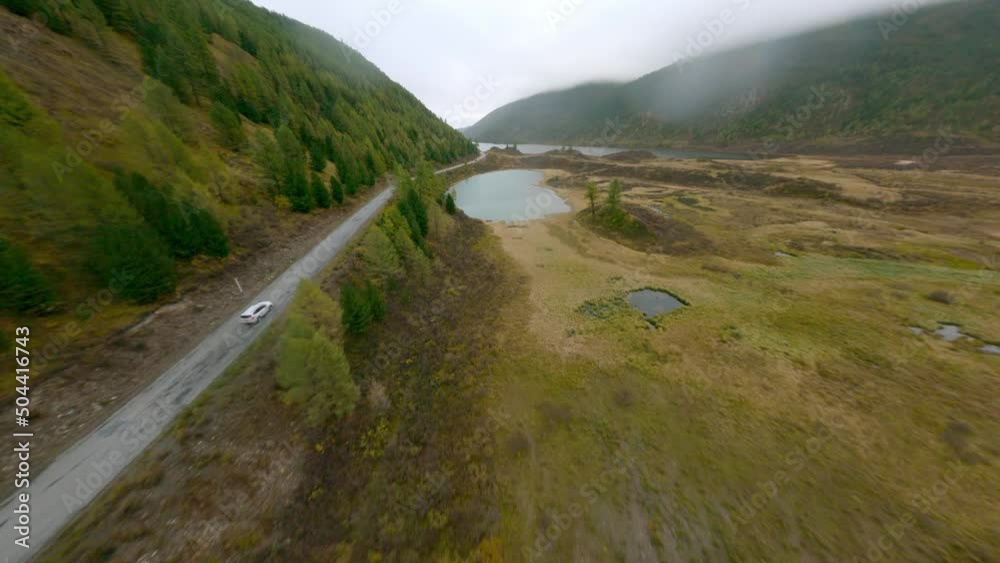 Aerial view white SUV automobile riding on dirt road near natural forest and mountain lake cloudscape rocky scenery. FPV sports drone flying over picturesque overcast weather valley hilly terrain Aerial view white SUV automobile riding on dirt road near natural forest and mountain lake cloudscape rocky scenery. FPV sports drone flying over picturesque overcast weather valley hilly terrain