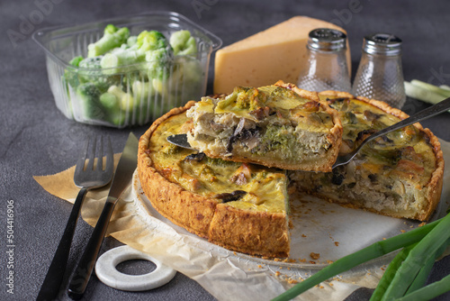 Quiche with chicken, mushrooms, broccoli and cheese, homemade traditional french pie with a cut piece on a dark gray background