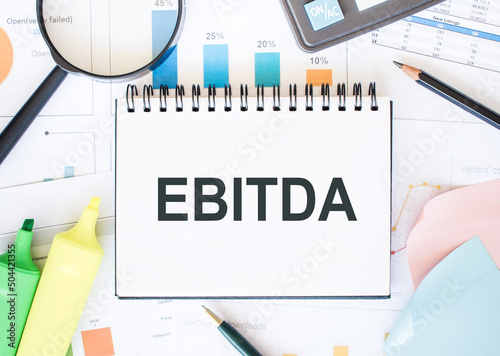 Text EBITDA on a notebook on diagram and charts with calculator, magnifier and pen
