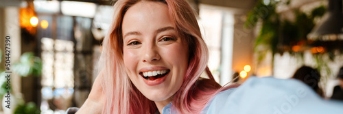 Young woman with pink hair smiling and taking selfie photo in cafe
