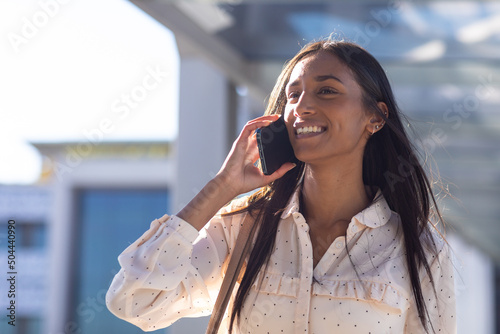 Beautiful smiling young biracial businesswoman talking through smartphone in corridor on sunny day