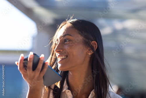 Happy young biracial businesswoman talking through smartphone speaker in corridor on sunny day