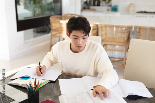 High angle view of asian teenage boy writing homework in book on table while sitting at home photo