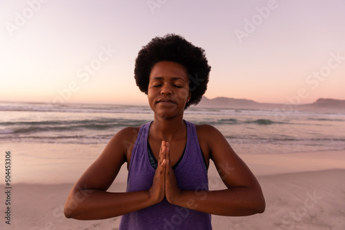 African american mature woman with afro hair meditating in prayer pose against sea and clear sky