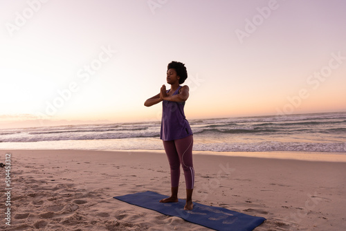 Full length of african american mature woman meditating in prayer pose on mat at beach at sunset